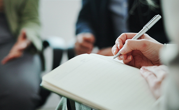 Person schreibt mit einem Stift in ein Notizbuch w&auml;hrend andere Personen im Hintergrund sitzen