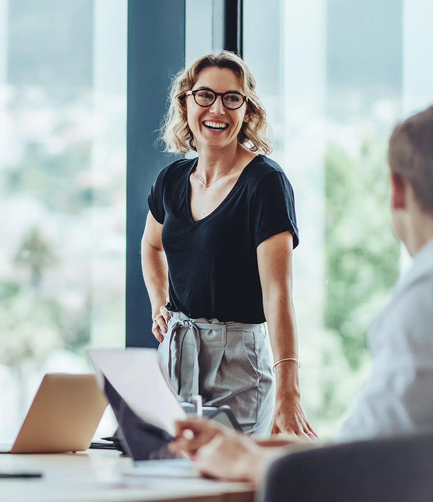 Selbstbewusste Frau steht am Tischende und lacht, w&auml;hrend andere zuh&ouml;ren